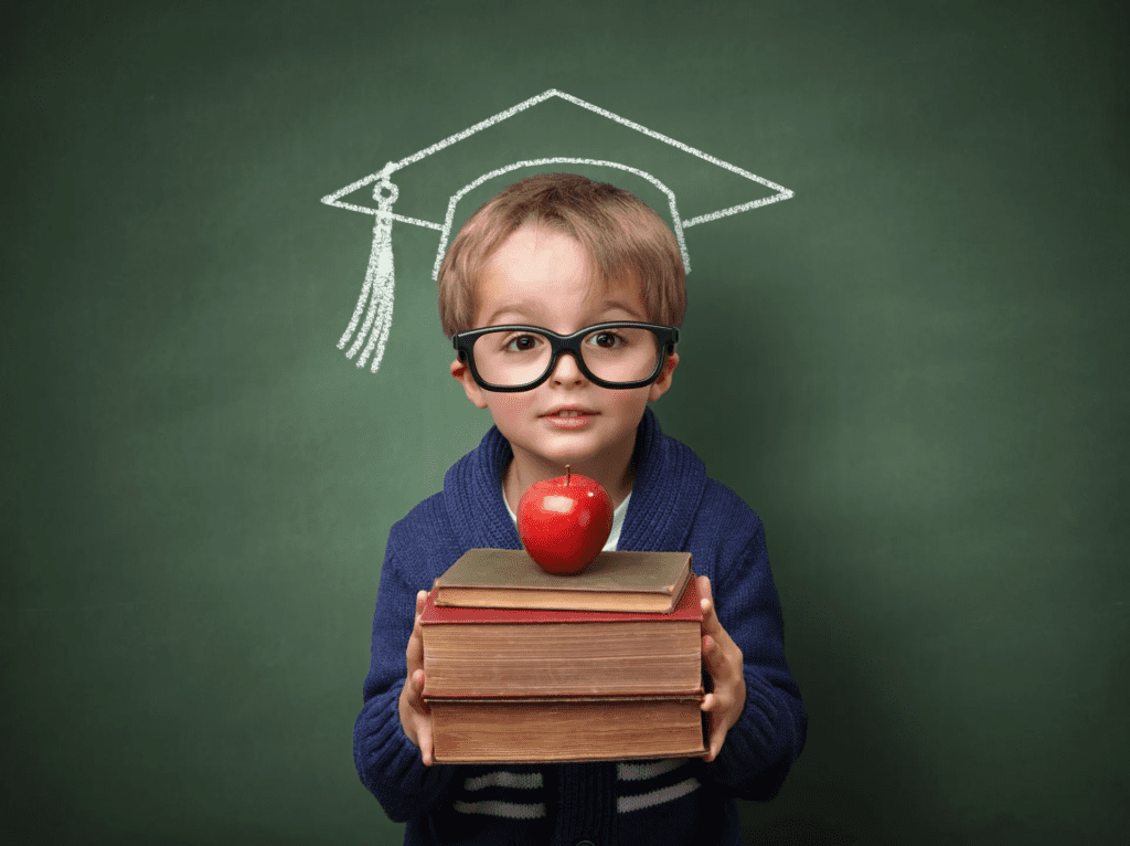 Little Blonde Boy Wearing Glasses and Holding a Stack of Books with an Apple on Top in Front of a Green Board with a Graduation Cap Drawn in Chalk