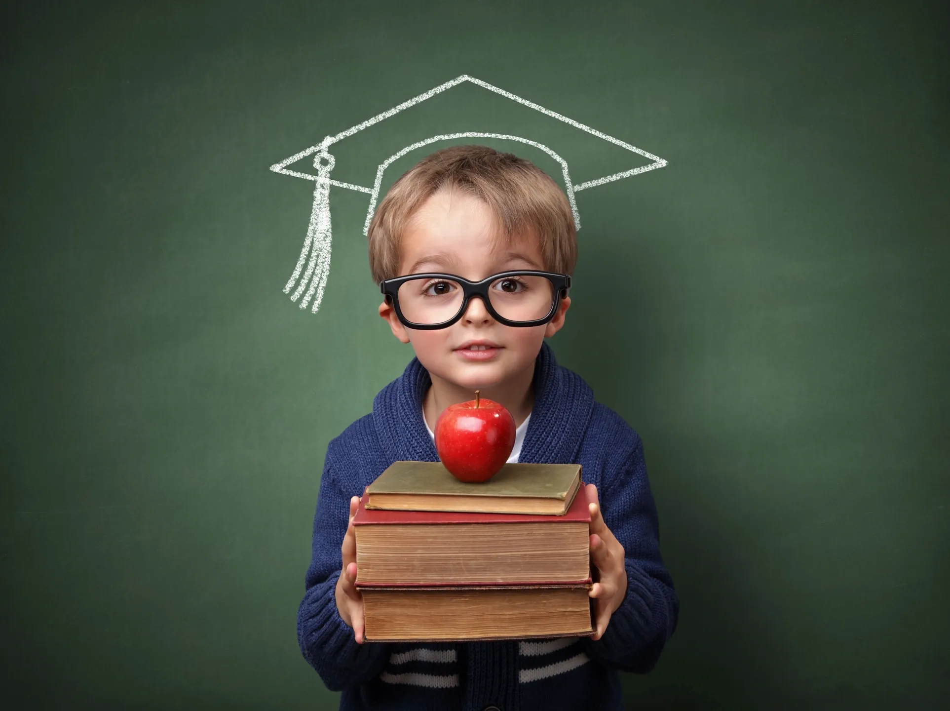 Little Blonde Boy Wearing Glasses and Holding a Stack of Books with an Apple on Top in Front of a Green Board with a Graduation Cap Drawn in Chalk