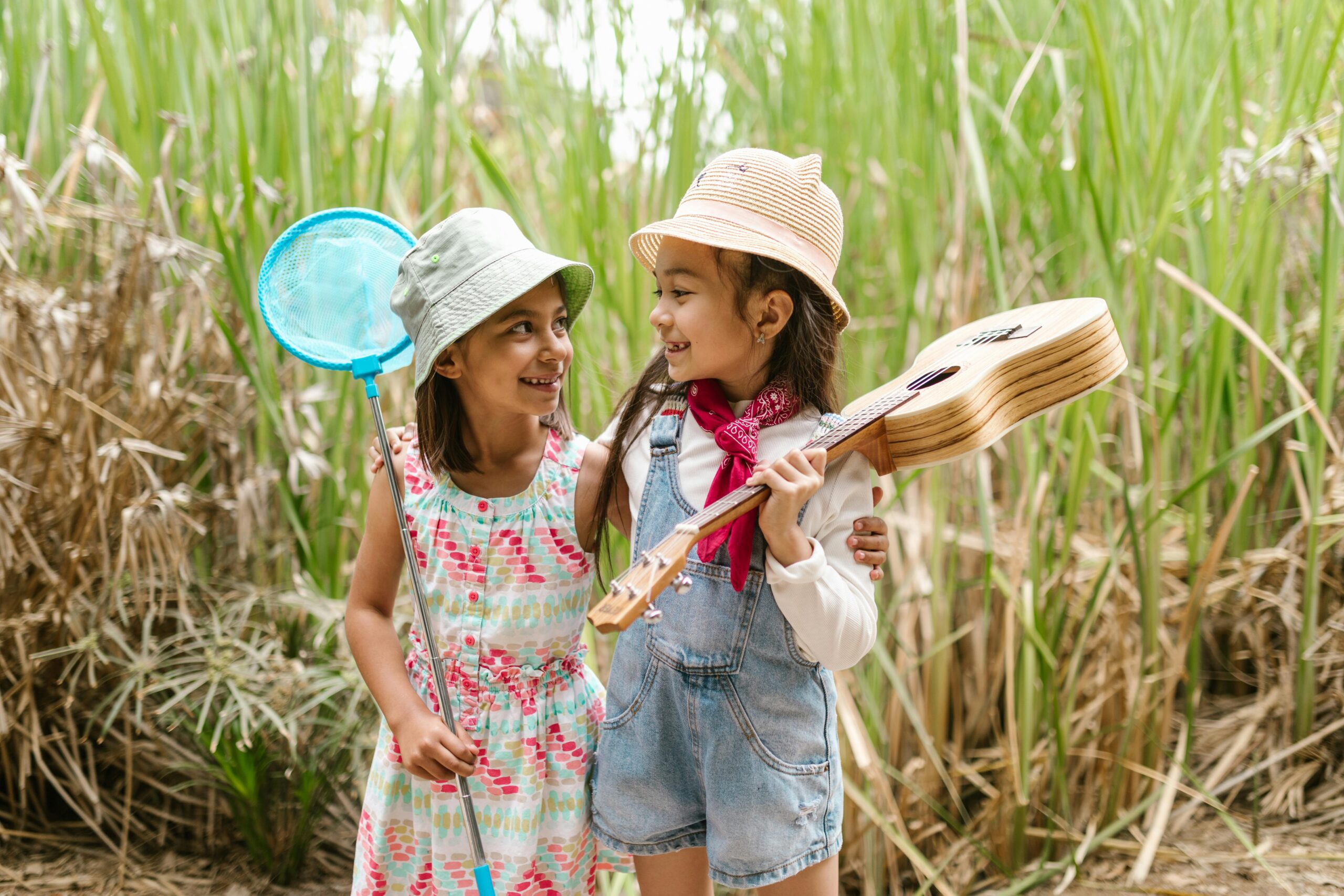 Two Young Girls in Nature Wearing Hats and Holding a Guitar and a Net