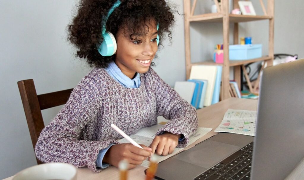 Teen Student Listening to a Course Using Headphones and Looking at a MacBook While Writing in a Notebook