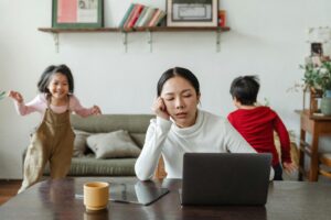 Asian mother looking tired at a computer with a girl and boy running around in the background